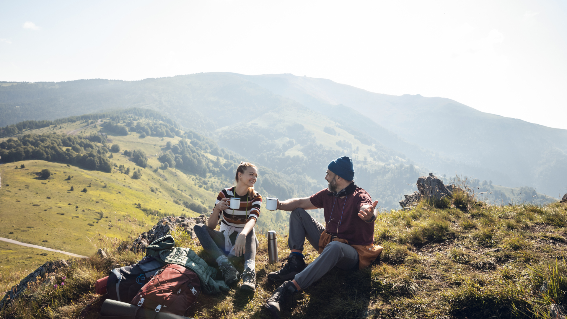 Un hombre y una mujer disfrutan de bebidas calientes mientras viajan con mochila con las colinas de fondo.jpg