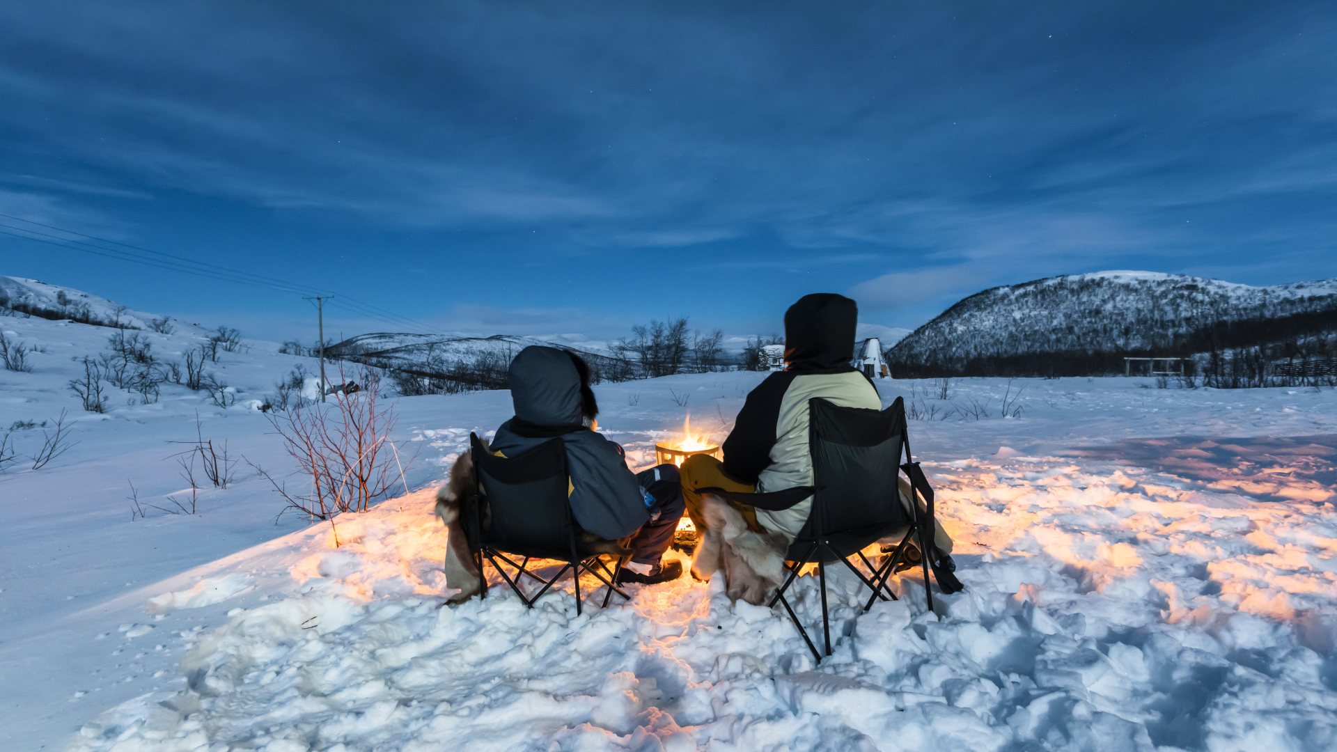 Dos personas se sientan frente a una fogata en la nieve.